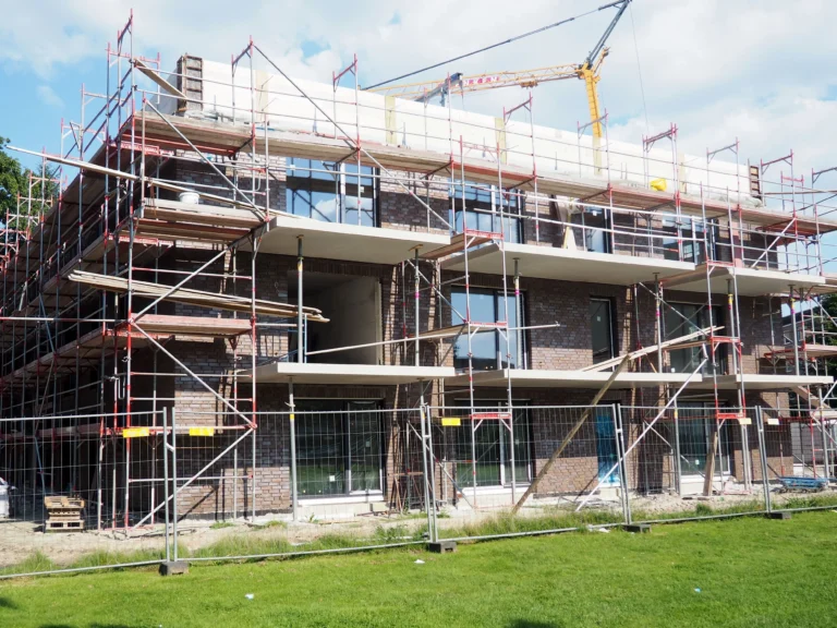Architekt Oldenburg: Baustelle eines Mehrfamilienhauses im Rohbau zwecks Neubaus. Eine rote Klinkerfassade mit Gerüst abgegrenzt ohne Dach von einem Bauzaun auf der grünen wiese mit Baukran im Hintergrund. Teilweise sind Fenster eingesetzt.