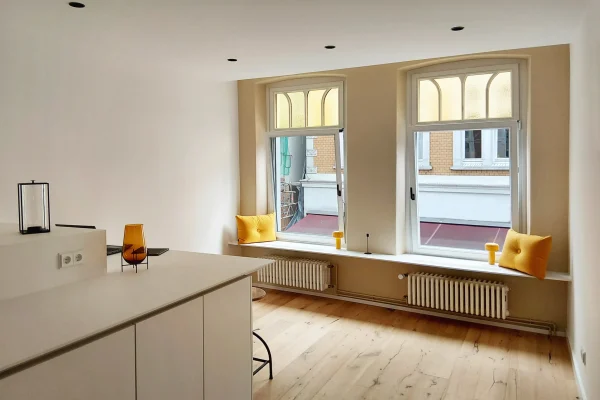 Ein heller, minimalistischer Küchen- und Wohnbereich mit hellen Holzböden, weißen Wänden und zwei großen Fenstern. Gelbe Kissen und Glasdekor bringen Farbe auf die Fensterplätze und den Tresen.