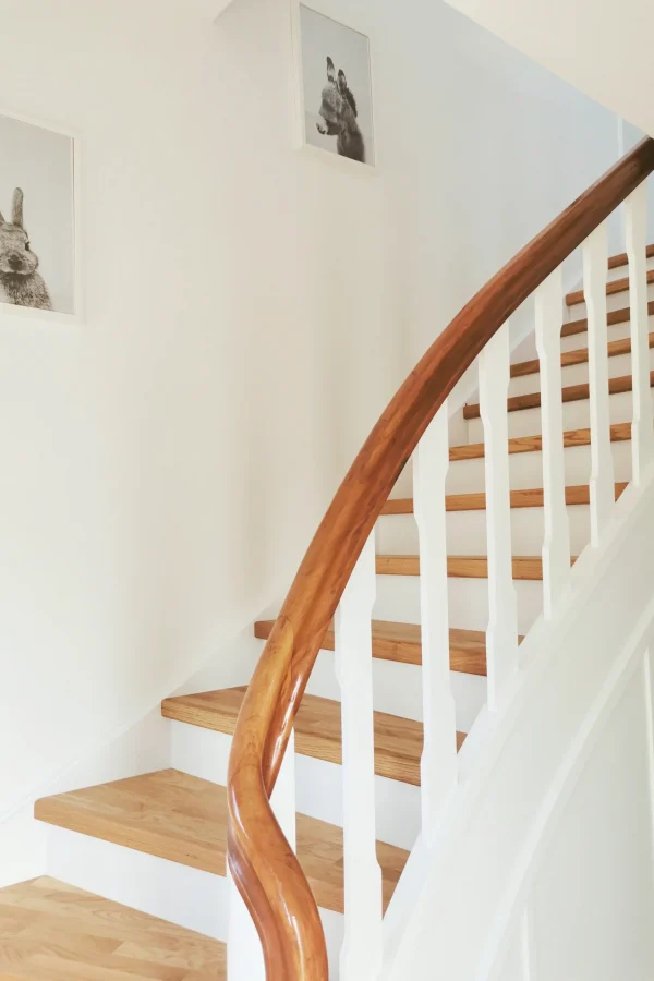 Eine Holztreppe mit weißen Setzstufen und einem polierten Holzhandlauf schlängelt sich nach oben. Zwei gerahmte Tierbilder, darunter ein Kaninchen, hängen an den weißen Wänden. Natürliches Licht erhellt den Raum.
