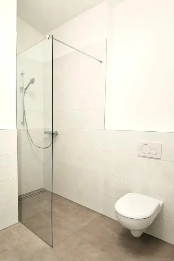Minimalistisches Badezimmer mit einer begehbaren Dusche, die von einer klaren Glaswand umschlossen ist, einer wandmontierten Kopfbrause und einer wandmontierten Toilette mit Zweimengenspülung an einer weiß gefliesten Wand. Der Fußboden ist hellbeige.