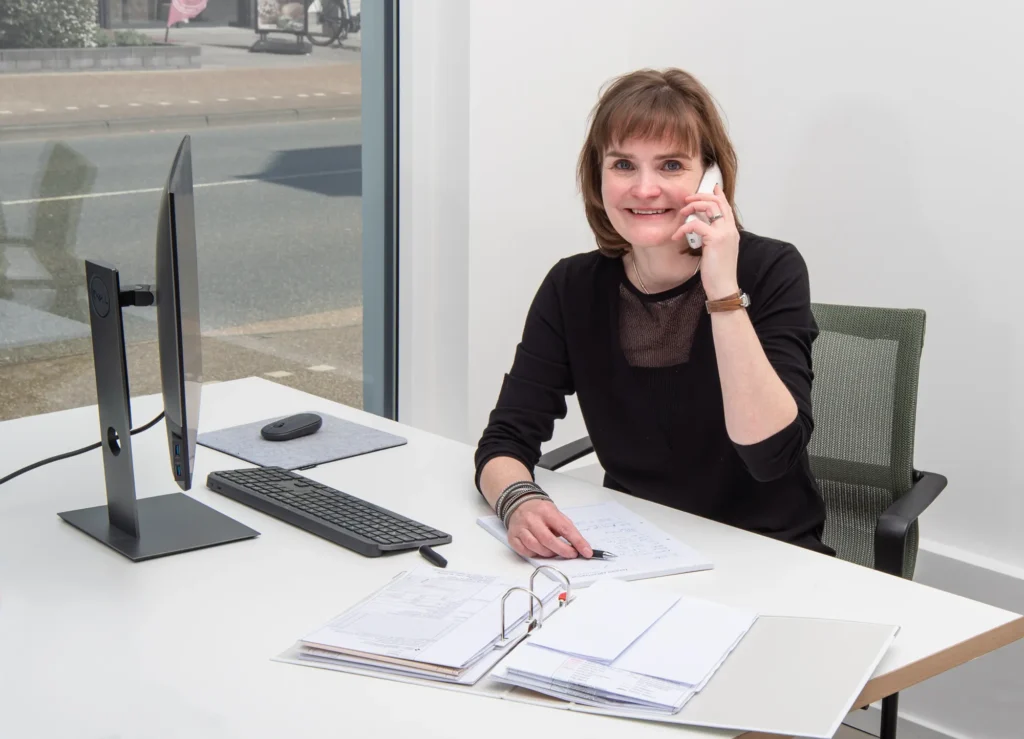 Eine Frau sitzt an einem Schreibtisch in einem modernen Architekturbüro in Oldenburg und lächelt, während sie in ein Festnetztelefon spricht. Offene Ordner, Papiere und ein Computer stehen auf ihrem Schreibtisch. Durch ein großes Fenster in der Nähe fällt natürliches Licht ein.