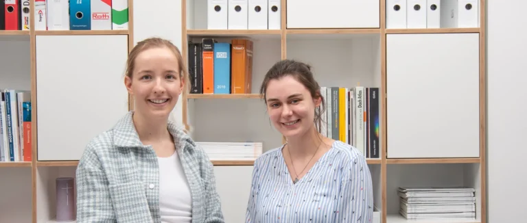 Zwei Frauen stehen lächelnd vor einem Bücherregal. Die Regale hinter ihnen sind mit Büchern, Akten und Zeitschriften gefüllt.