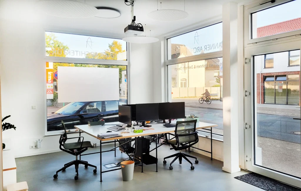 Ein modernes Büro mit großen Fenstern, zwei Schreibtischen mit Computern und Stühlen, einem Projektor und einer Straßenansicht, die ein Auto und einen Radfahrer zeigt. Das Sonnenlicht erhellt den sauberen, minimalistischen Arbeitsbereich.