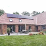 Ein Backsteinhaus mit rotem Ziegeldach und Sonnenkollektoren, mit großen Glastüren, Terrassenmöbeln, Topfpflanzen und einem grünen Rasen im Vordergrund. Im Hintergrund sind Bäume zu sehen.