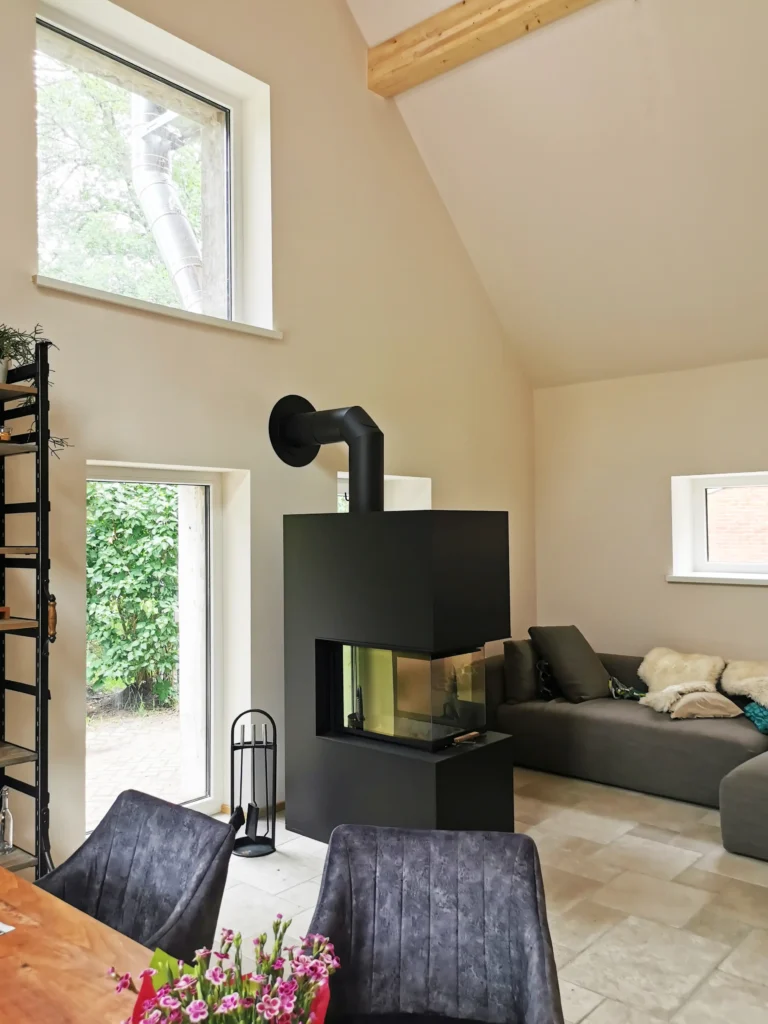 Ein modernes Wohnzimmer, das von Architekten mit Erfahrung in der Altbausanierung gestaltet wurde, mit großen Fenstern, einem schwarzen Holzofen, einem grauen Sektionssofa mit Decken, Fliesenboden und einem Esstisch mit Blumen im Vordergrund.