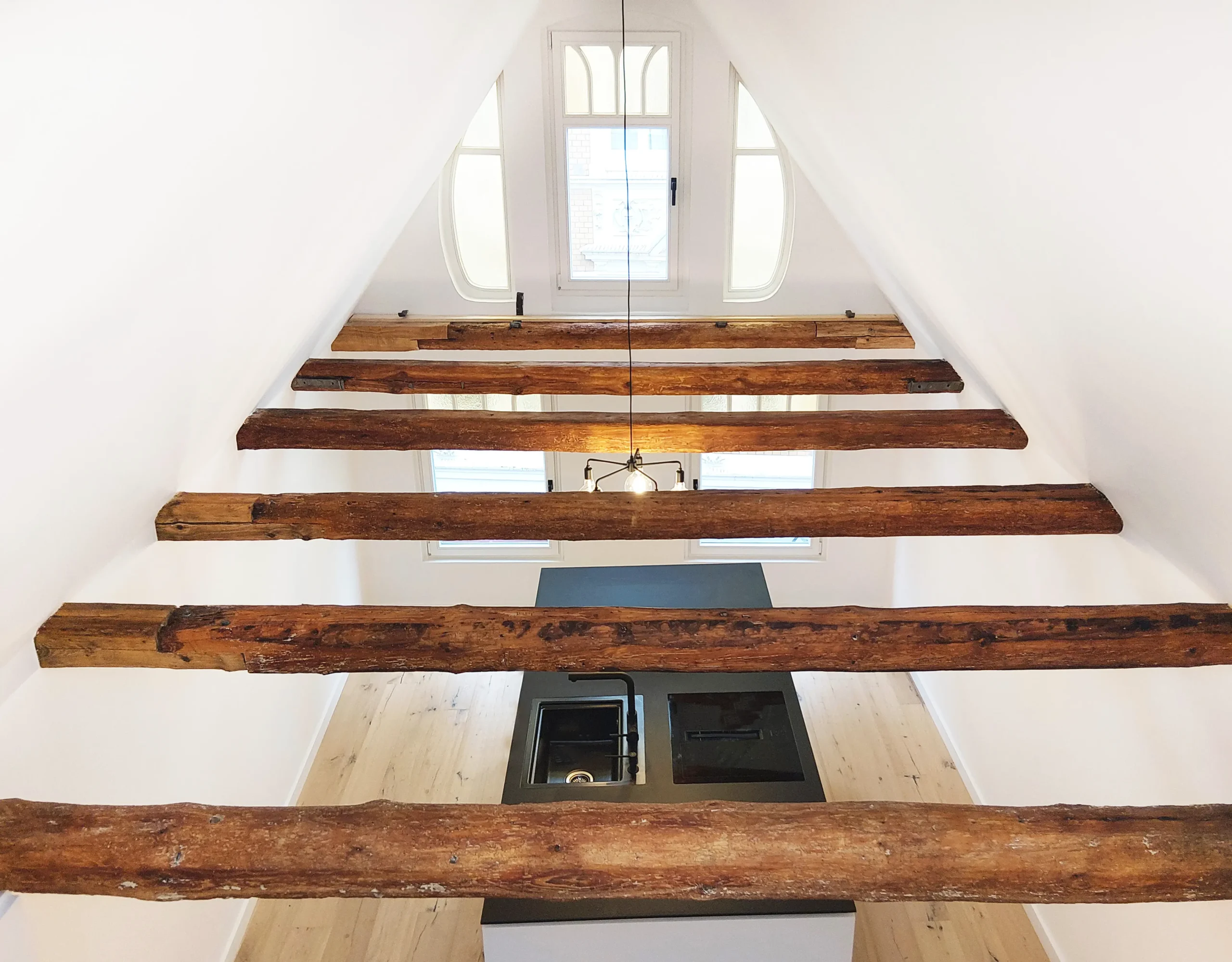 Eine helle Dachgeschossküche mit freiliegenden Holzbalken, hellen Holzböden, einer zentralen Insel mit Spüle und Herd und Bogenfenstern, die viel Tageslicht hereinlassen - entworfen von einem Oldenburger Architekten.