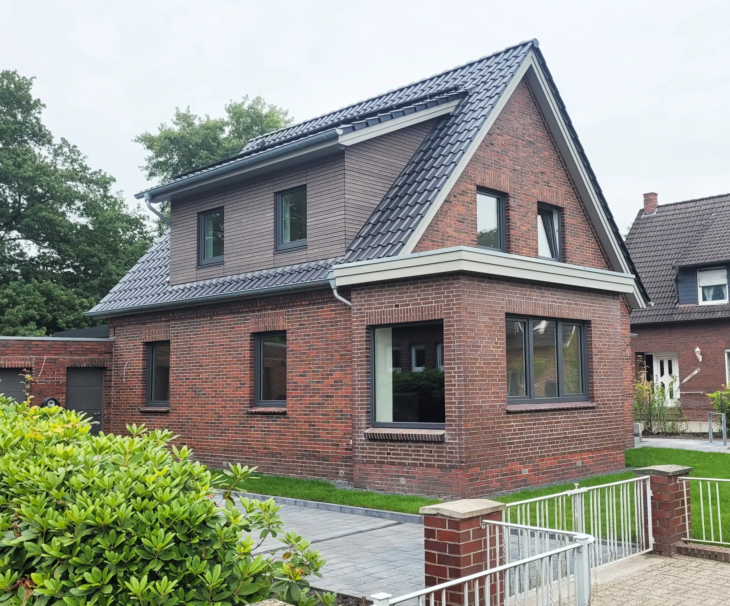 Ein modernes Backsteinhaus mit einem steilen, dunkelgrauen Ziegeldach und großen Fenstern, umgeben von einer kleinen Rasenfläche, einem Gehweg und einem weißen Metallzaun. Im Hintergrund sind Büsche und benachbarte Häuser zu sehen.
