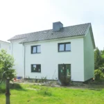 Ein zweistöckiges weißes Haus mit einem dunklen Ziegeldach steht in einem grünen Garten mit Sträuchern, Bäumen und lila Blumen. Das Haus hat mehrere Fenster und eine Tür, und der Hof ist sonnig und grasbewachsen.