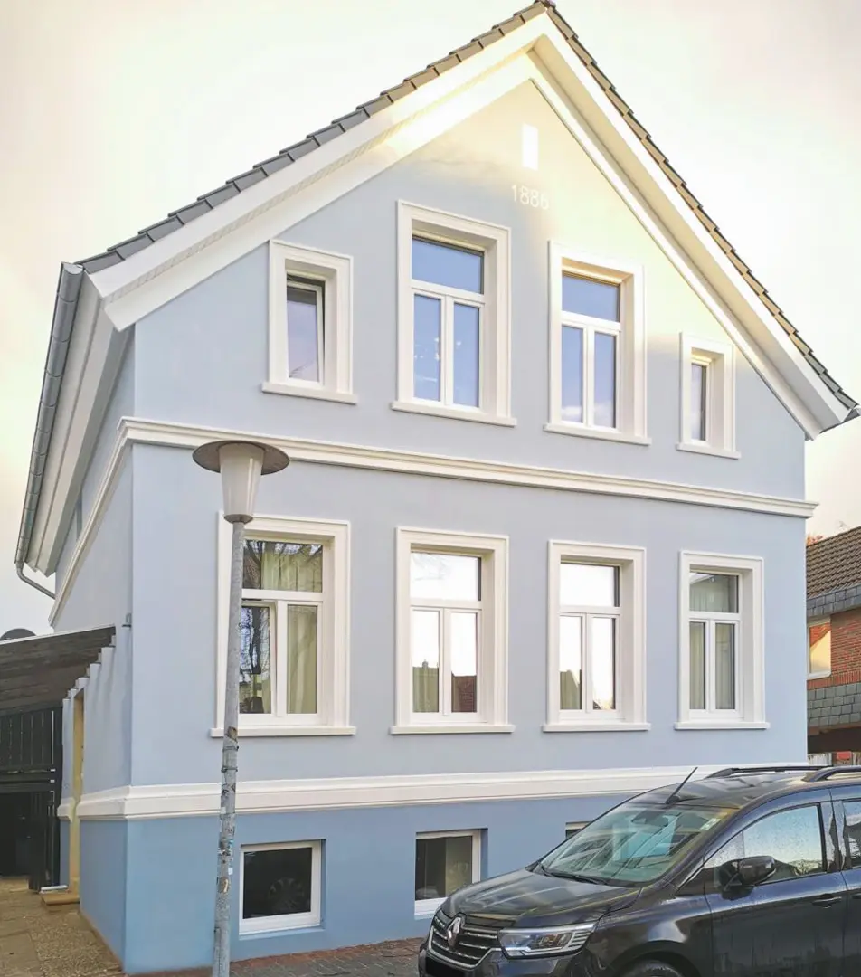 Ein hellblaues, zweistöckiges Haus mit weißen Zierleisten, mehreren Fenstern und einem schrägen Dach. Davor steht eine Straßenlaterne, und neben dem Haus ist ein schwarzes Auto geparkt. Die Jahreszahl 1898 ist in der Nähe der Dachspitze sichtbar.