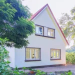 Ein weißes Haus mit rotem Ziegeldach und großen Fenstern, umgeben von grünen Bäumen und Sträuchern, mit einer gepflasterten Fläche im Vordergrund.