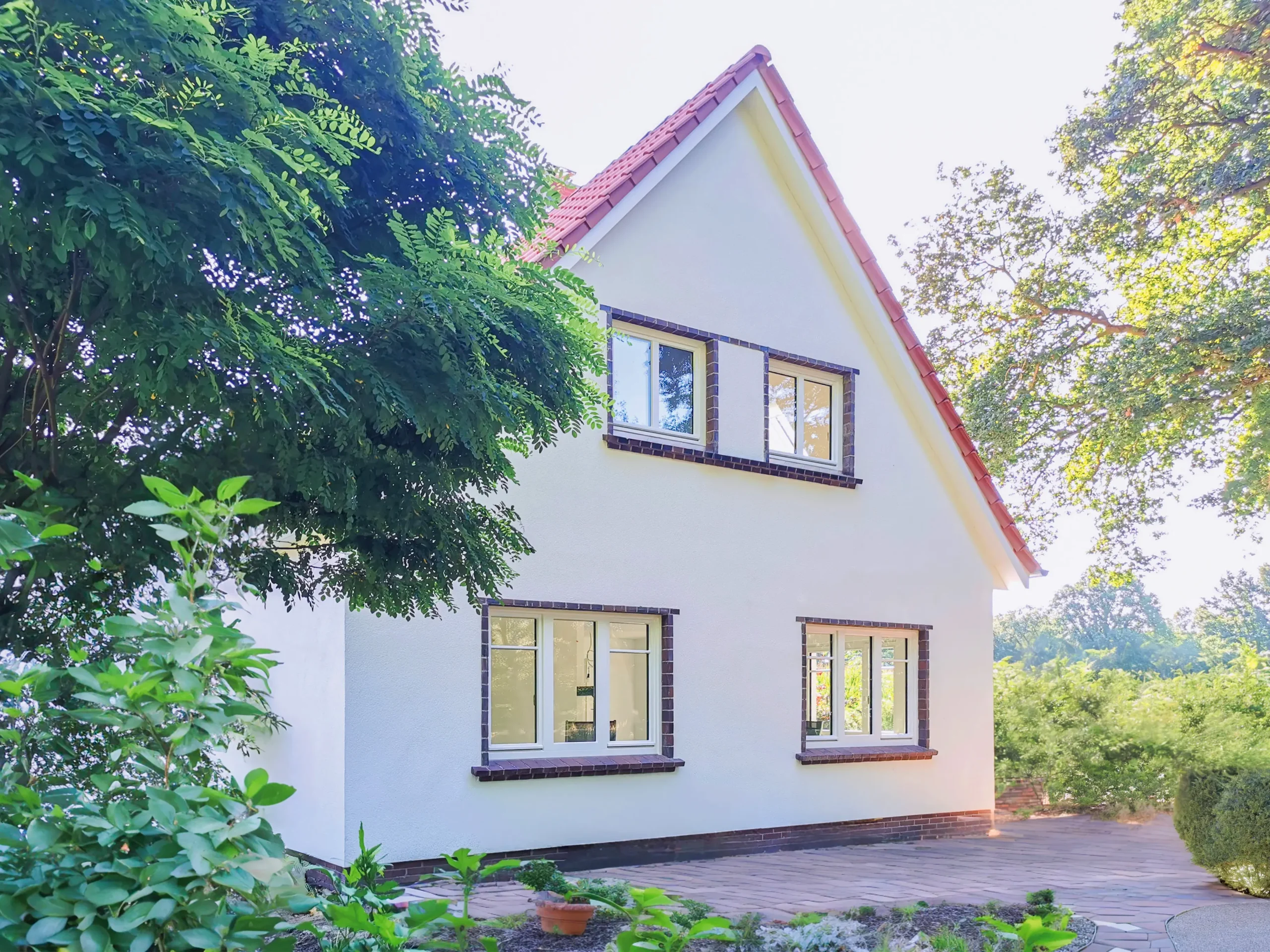 Ein weißes Haus mit rotem Ziegeldach und großen Fenstern, umgeben von grünen Bäumen und Sträuchern, mit einer gepflasterten Fläche im Vordergrund.