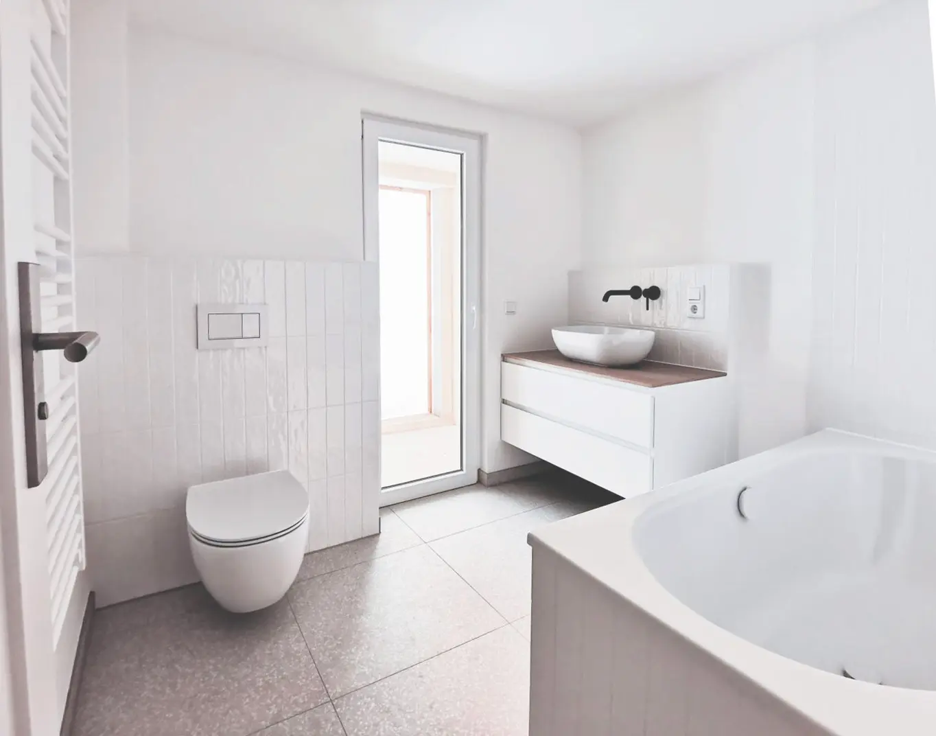 Modernes, minimalistisches Badezimmer, das von einem Architekten für Umbau entworfen wurde. Es besteht aus einer Wandtoilette, einem freistehenden Waschbecken auf einem weißen Waschtisch, einer Badewanne, einem gefliesten Boden und einer Tür, die in einen sonnendurchfluteten Bereich führt. Der Raum ist hell und überwiegend weiß.