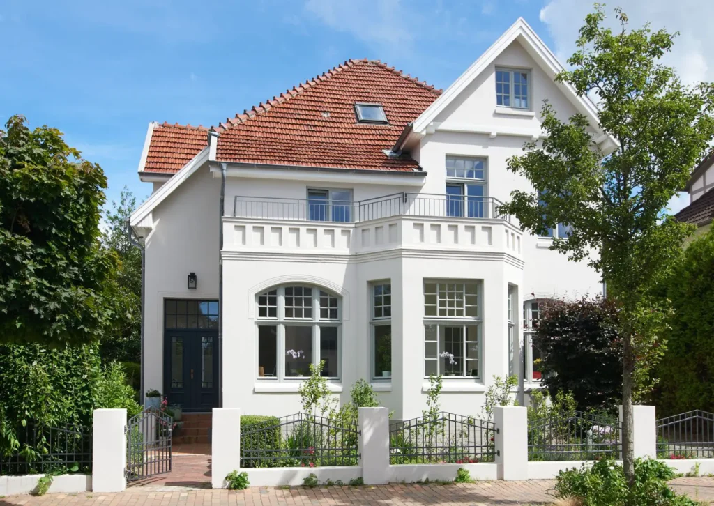 Ein zweistöckiges weißes Haus mit rotem Ziegeldach, einem Balkon, großen Fenstern und viel Grün, hinter einem niedrigen weißen Zaun und einem gemauerten Weg gelegen.