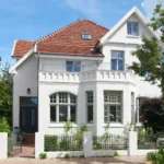 Ein zweistöckiges weißes Haus mit rotem Ziegeldach, einem Balkon, großen Fenstern und viel Grün, hinter einem niedrigen weißen Zaun und einem gemauerten Weg gelegen.