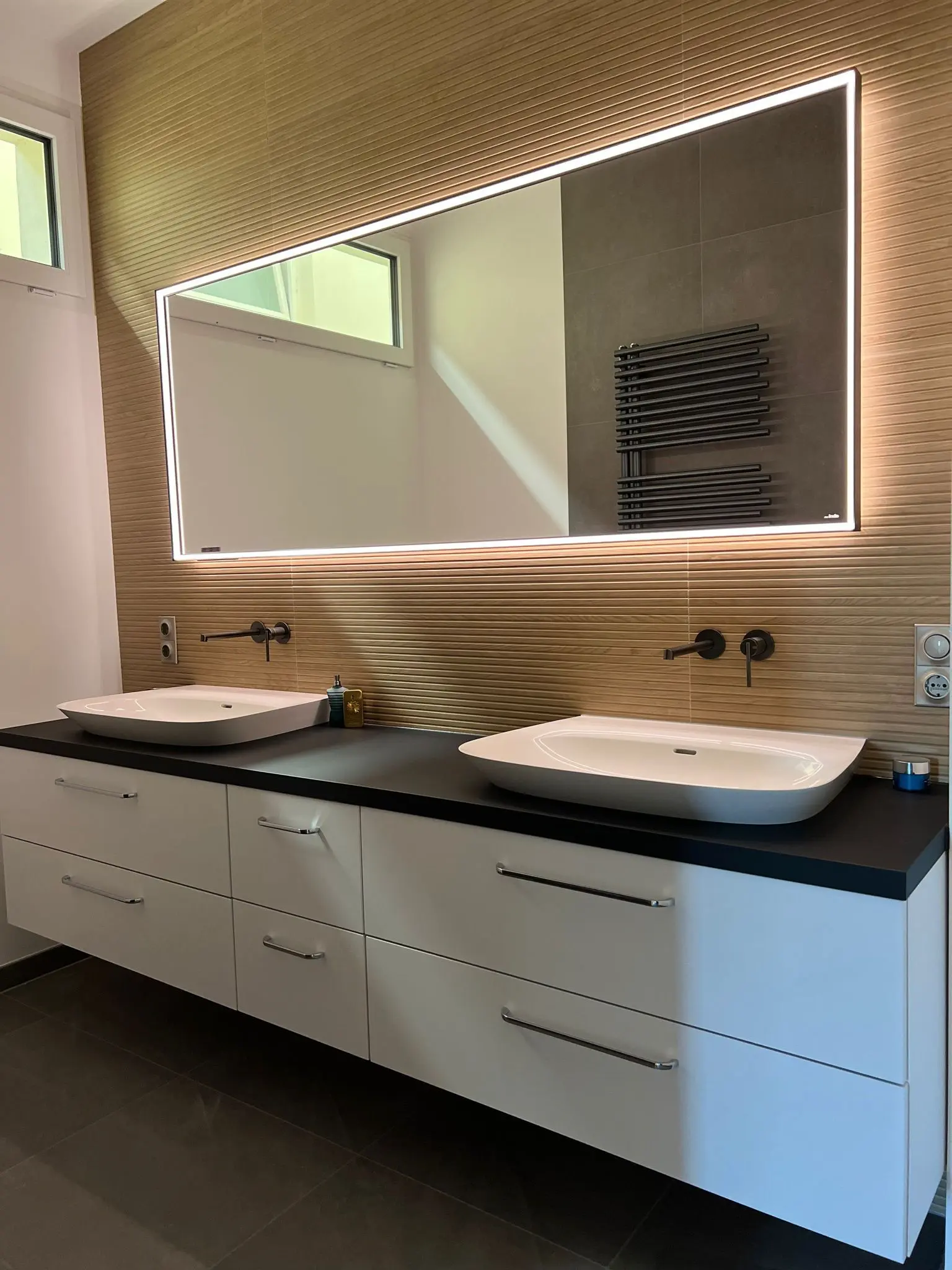 Modernes Badezimmer mit einem großen, beleuchteten rechteckigen Spiegel über einer schwarzen Arbeitsplatte mit zwei weißen Waschbecken und Unterbau-Schubladen; Handtuchwärmer und hohe Fenster im Hintergrund.