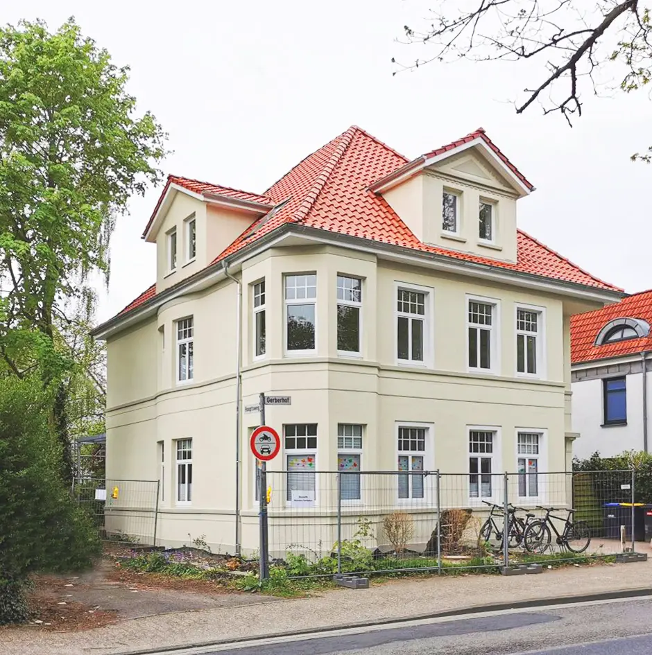 Ein zweistöckiges cremefarbenes Haus mit rotem Ziegeldach, umgeben von einem Zaun. Es gibt mehrere Fenster, zwei Fahrräder am Zaun und Bäume in der Nähe. Vor dem Haus ist ein Schild "Betreten verboten" zu sehen.