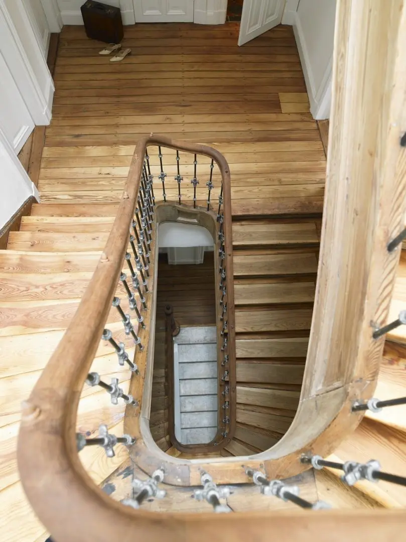 Blick vom oberen Ende einer von einem Oldenburger Architekten entworfenen Holztreppe mit geschwungenem Handlauf und schwarzen Spindeln über mehrere Stockwerke hinunter zu einer unteren Ebene mit helleren Stufen und Holzböden.