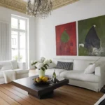 Ein modernes Wohnzimmer mit weißen Sofas, einem dunklen Couchtisch aus Holz mit Blumen, Obst und Wein, großen Fenstern, Holzböden und zwei abstrakten Gemälden an der Wand. Ein Kronleuchter hängt von einer verzierten Decke.