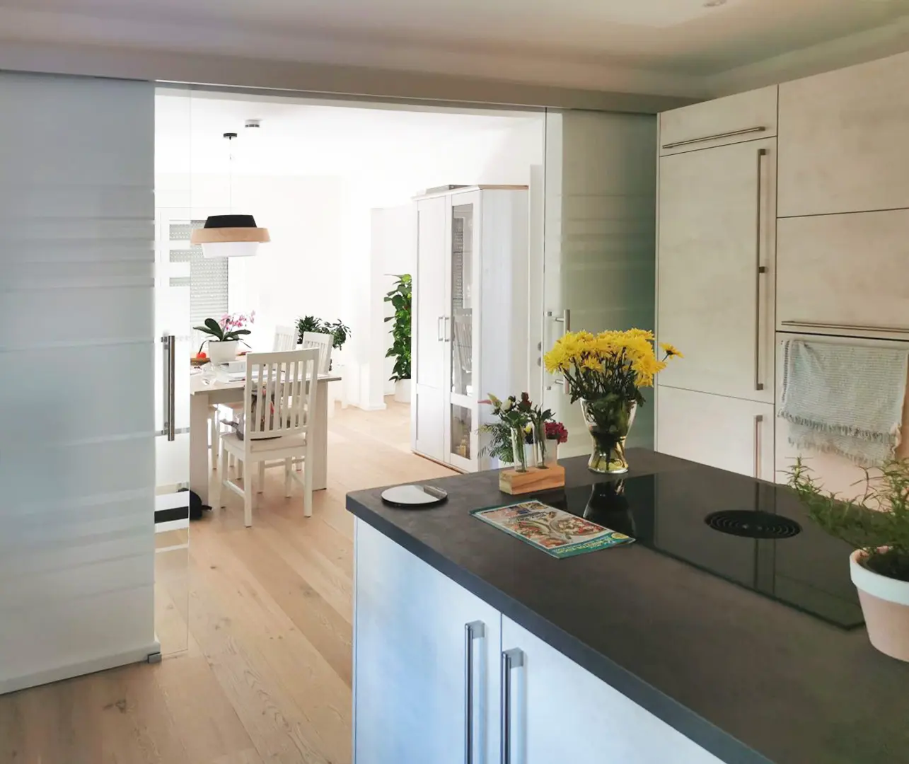 Eine moderne Küche mit hellem Holzboden, weißen Schränken und einer dunklen Arbeitsplatte. Gelbe Blumen in einer Vase, ein Kochbuch und Topfpflanzen schmücken die Kücheninsel. Im Hintergrund ist ein heller Essbereich zu sehen.
