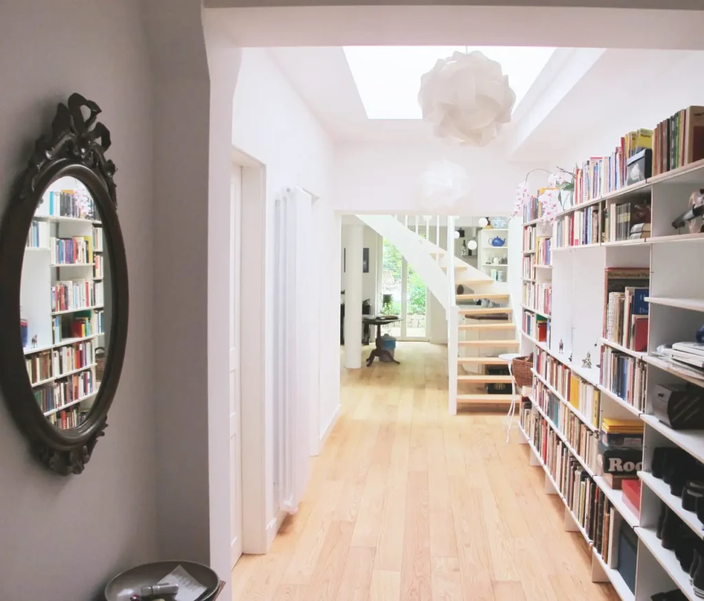 Ein heller Flur mit Holzfußboden, der rechts von Bücherregalen und links von einem dekorativen Spiegel gesäumt wird, führt zu einer Treppe und einem offenen Raum mit einem Schreibtisch und einem großen Fenster, das Tageslicht hereinlässt.