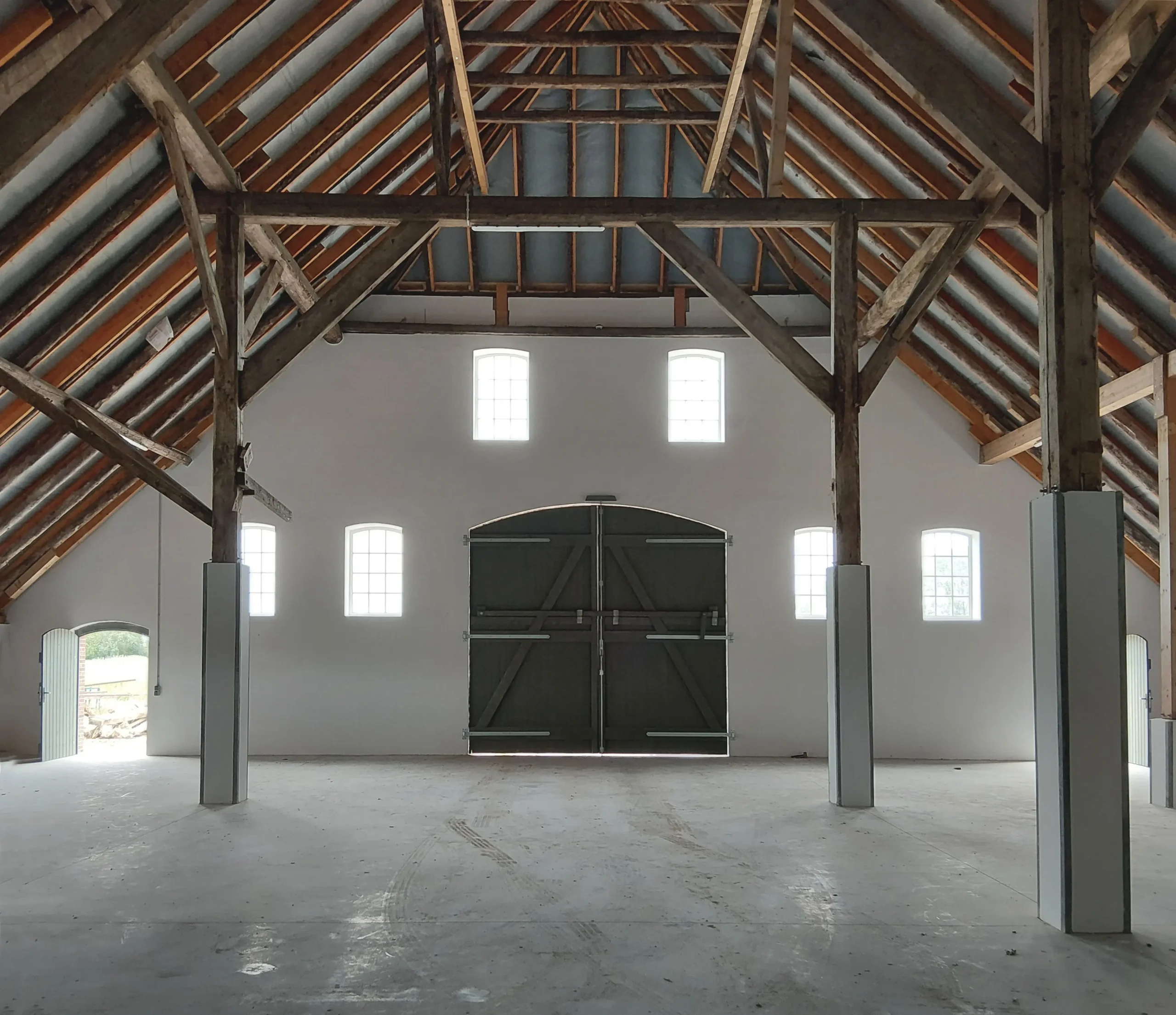 Das Innere einer geräumigen, leeren Scheune mit hoher Holzbalkendecke, weißen Wänden, großen Flügeltüren in der Mitte und mehreren hohen Fenstern, die Tageslicht hereinlassen.