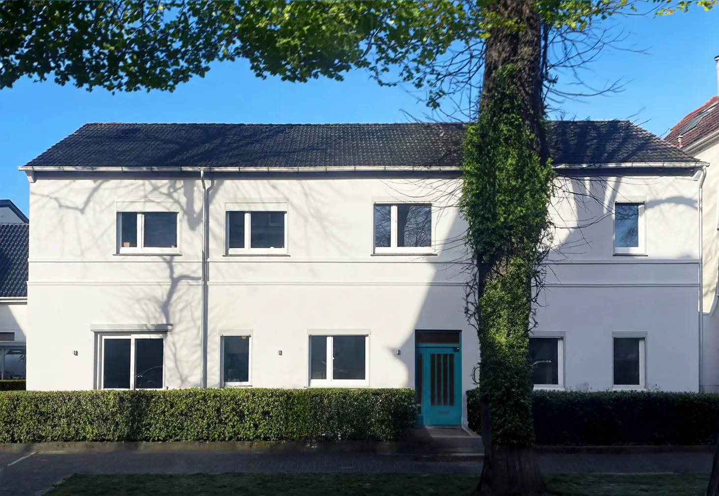 Ein zweistöckiges weißes Haus mit einem dunklen Ziegeldach, mehreren Fenstern, einer türkisfarbenen Eingangstür und einem Baum mit Efeu davor, unter einem klaren blauen Himmel.
