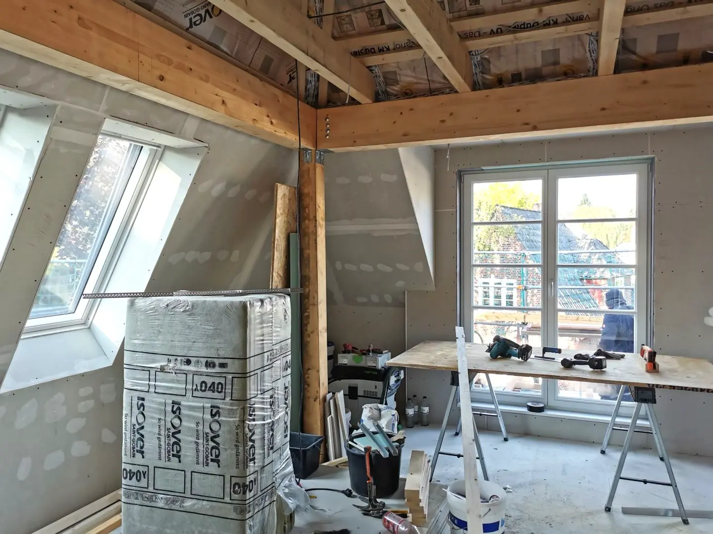 Ein Raum mit freiliegenden Balken, unfertigem Trockenbau, Werkzeugen, Baumaterialien und Dämmung. Sonnenlicht strömt durch große Fenster und ein Oberlicht herein und erhellt den Renovierungsraum.