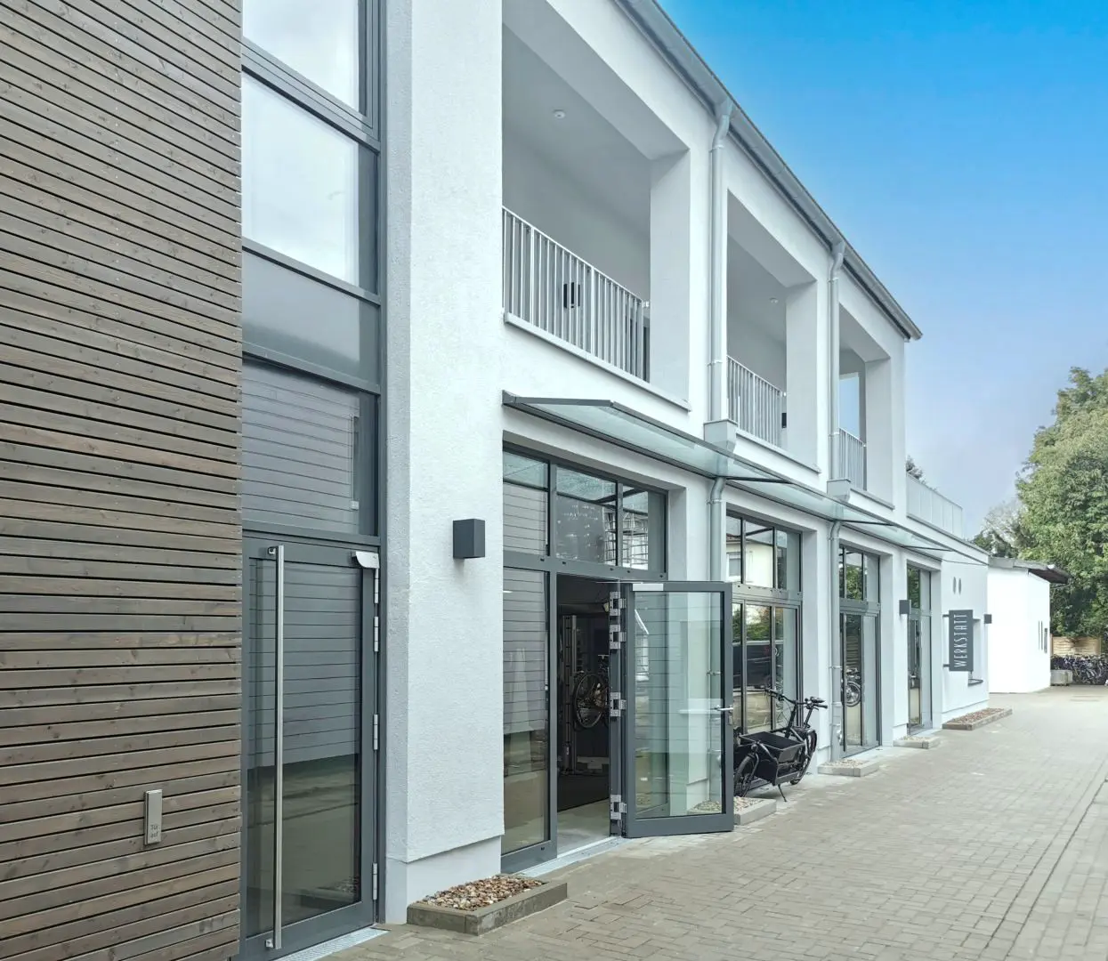 Modernes zweistöckiges Gebäude mit großen Fenstern und Glastüren, weißen Wänden und einem holzgetäfelten Bereich auf der linken Seite. Das von einem Oldenburger Architekten entworfene Gebäude ist mit einem Fahrrad ausgestattet, das draußen auf einem gepflasterten Gehweg unter blauem Himmel abgestellt ist.