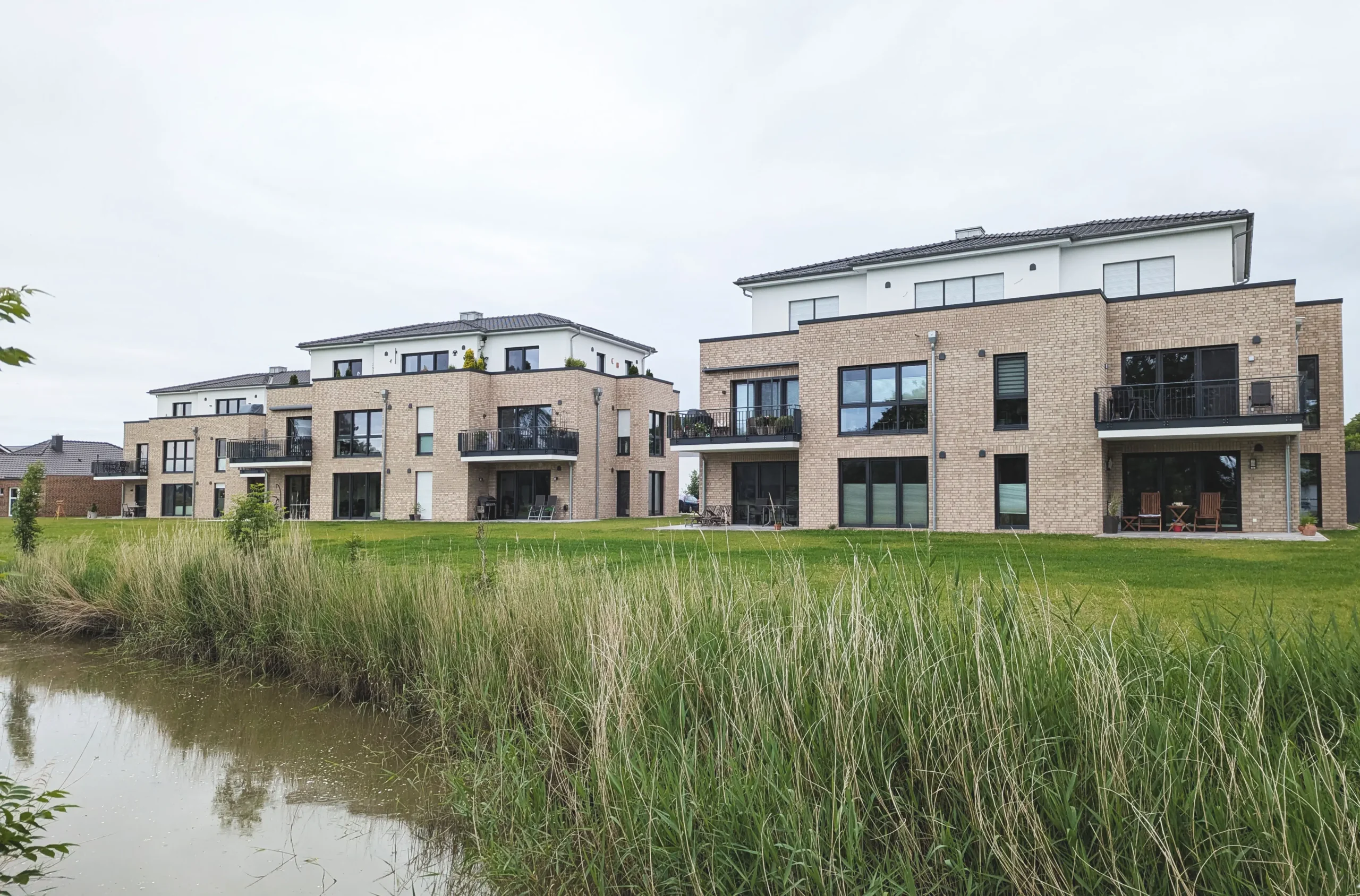Moderne, von einem Architekten für Neubau entworfene Wohnhäuser mit großen Fenstern und Balkonen stehen auf einer Rasenfläche in der Nähe eines kleinen, von hohem Gras gesäumten Teichs unter einem bewölkten Himmel.