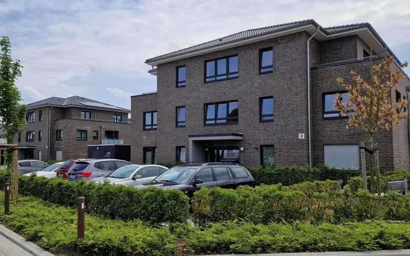 Ein modernes Backsteinwohnhaus mit großen Fenstern, entworfen von einem Architekten für Neubau, ist von Grün umgeben. Mehrere geparkte Autos stehen vor dem Gebäude unter einem teilweise bewölkten Himmel.