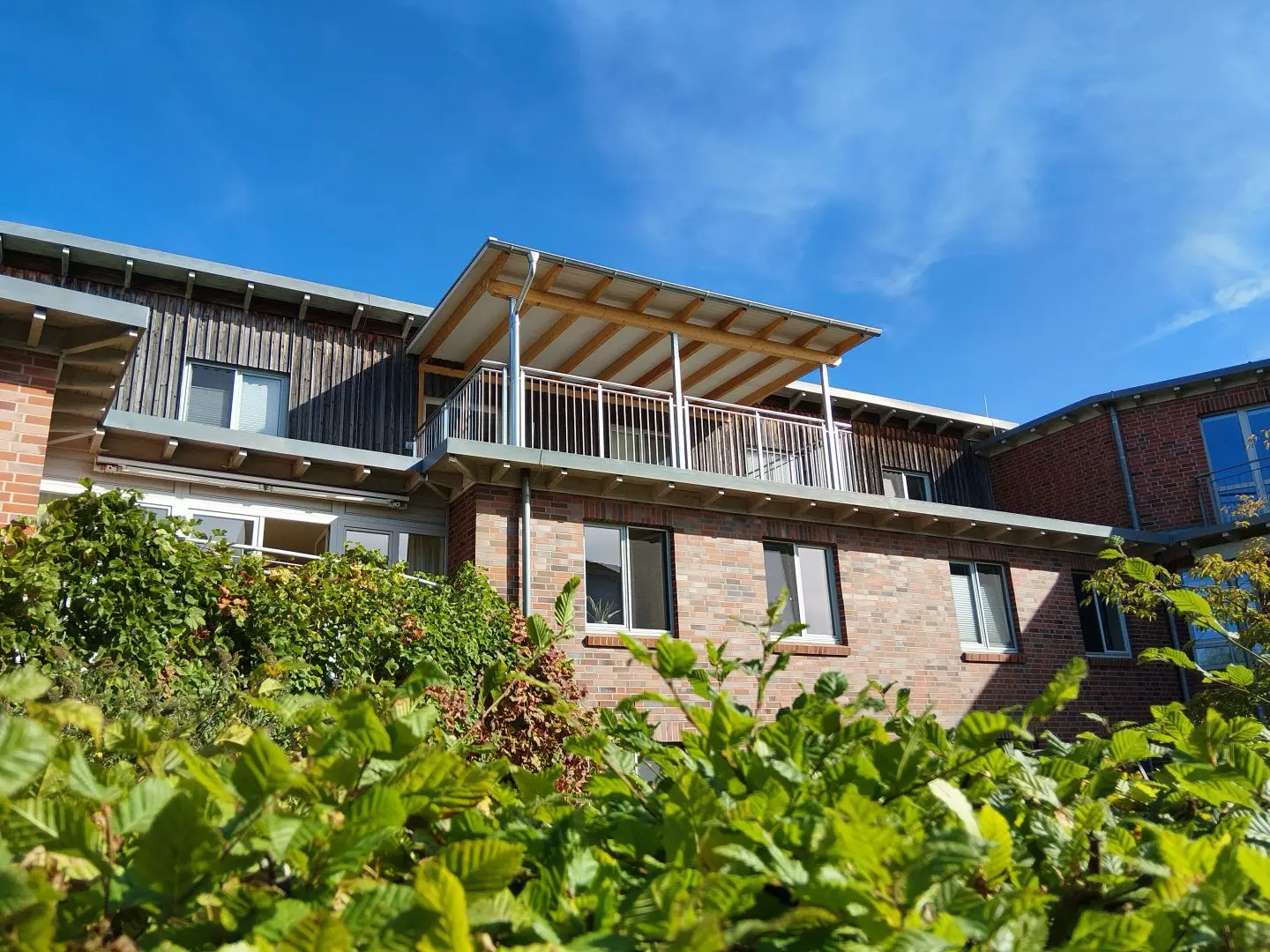 Ein zweistöckiges Backsteingebäude mit einem überdachten Balkon und großen Fenstern, umgeben von grünen Büschen, unter einem strahlend blauen Himmel mit Wolkenfetzen.
