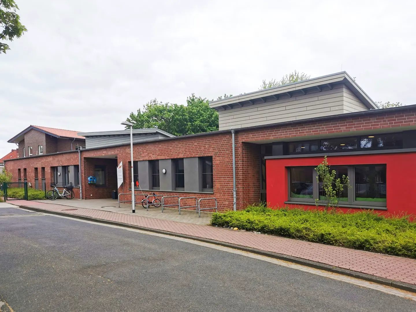 Ein modernes Backsteingebäude mit großen Fenstern, einem kleinen Fahrradständer und einem gepflasterten Gehweg davor. Draußen sind ein paar Fahrräder abgestellt, und das Gebäude ist bei bewölktem Himmel von Grün umgeben.